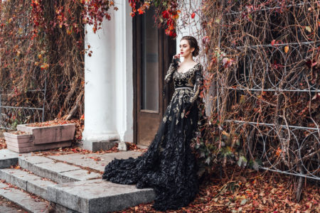Attractive young caucasian woman in black evening long dress posing near entrance of old house entwined with red autumn foliage